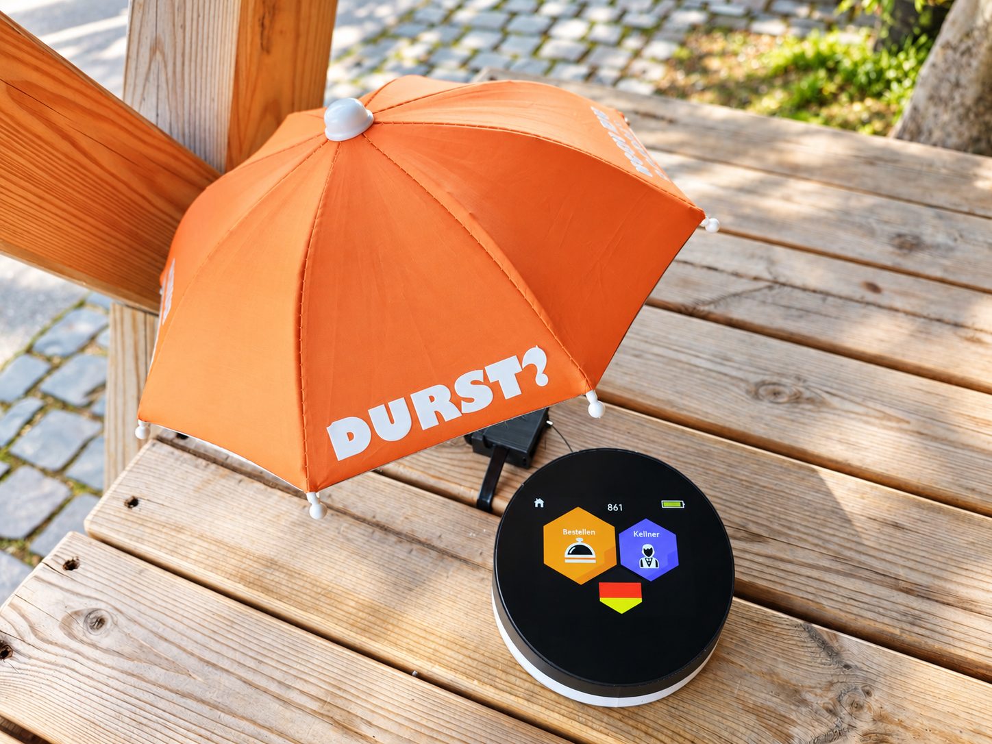 OrdiBee auf einem Holztisch: rundes Gerät mit Display, daneben ein orangefarbener Bestellschirm mit dem Spruch „DURST?“